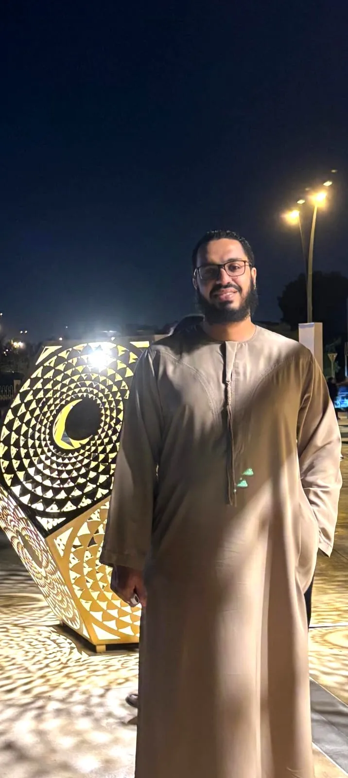 Mohamed in a kandura at an evening event in Dubai, standing beside a lit geometric lantern